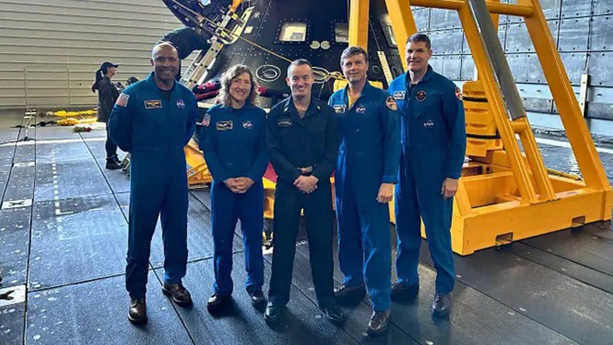 Five individuals posing in a row wearing blue NASA flight suits.